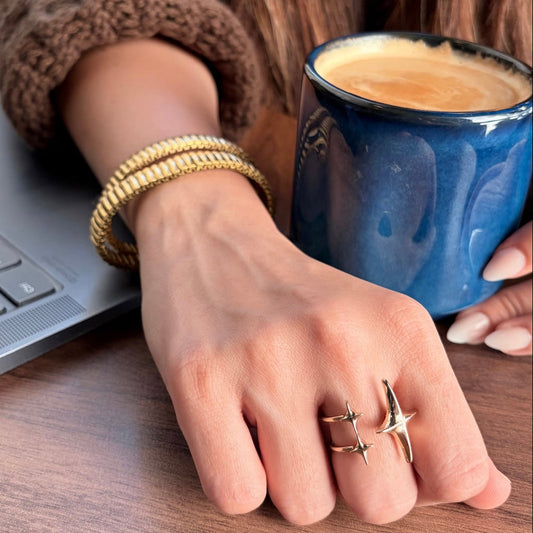 Hand with gold rings holding a blue mug of coffee next to a laptop on a wooden table